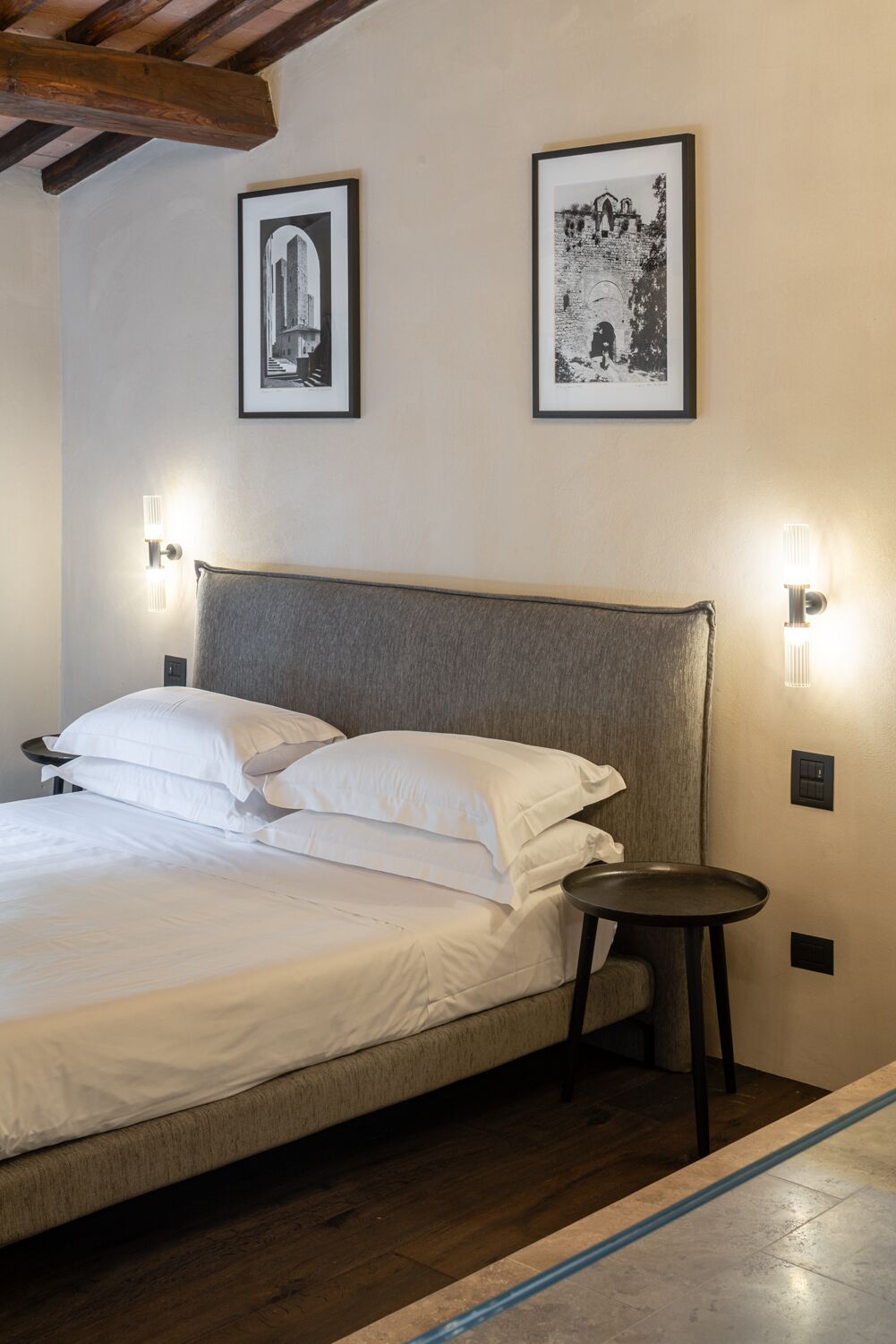 Una cama bien tendida con sábanas blancas, un cabecero tapizado en gris y dos fotografías en blanco y negro enmarcadas en la pared.