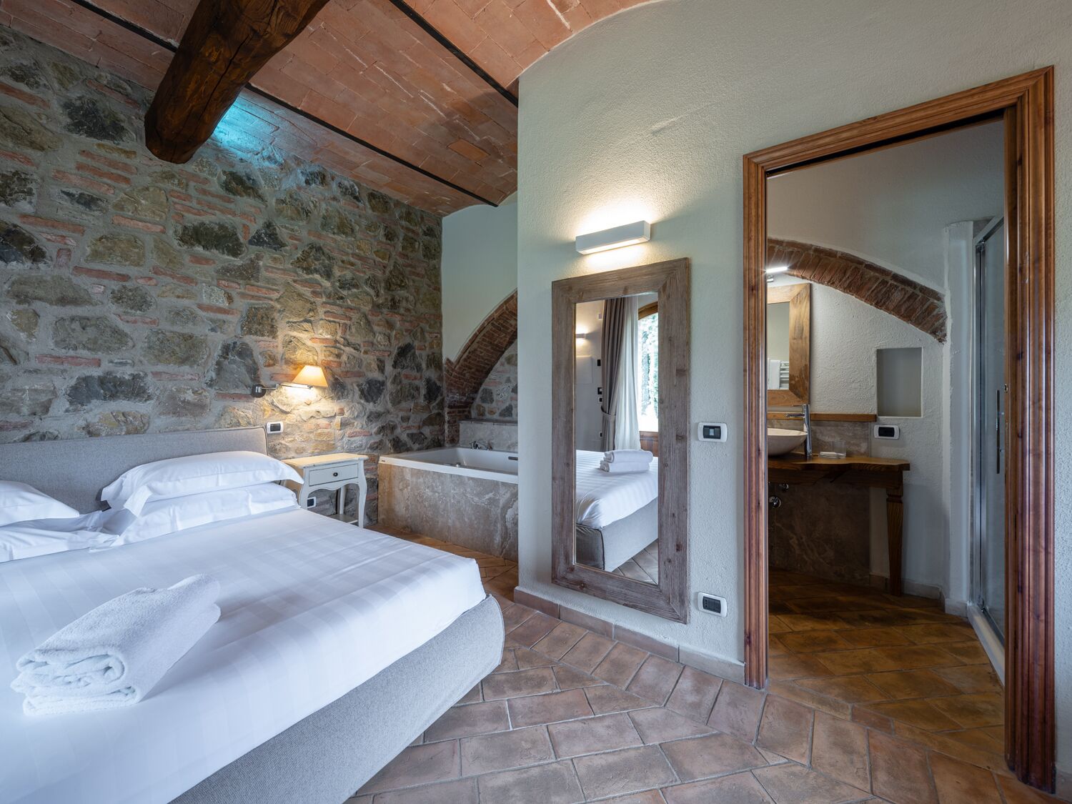 A bedroom with a stone wall, a wooden ceiling, a large bed, a bedside table, a mirror, and a bathroom with a bathtub and sink.