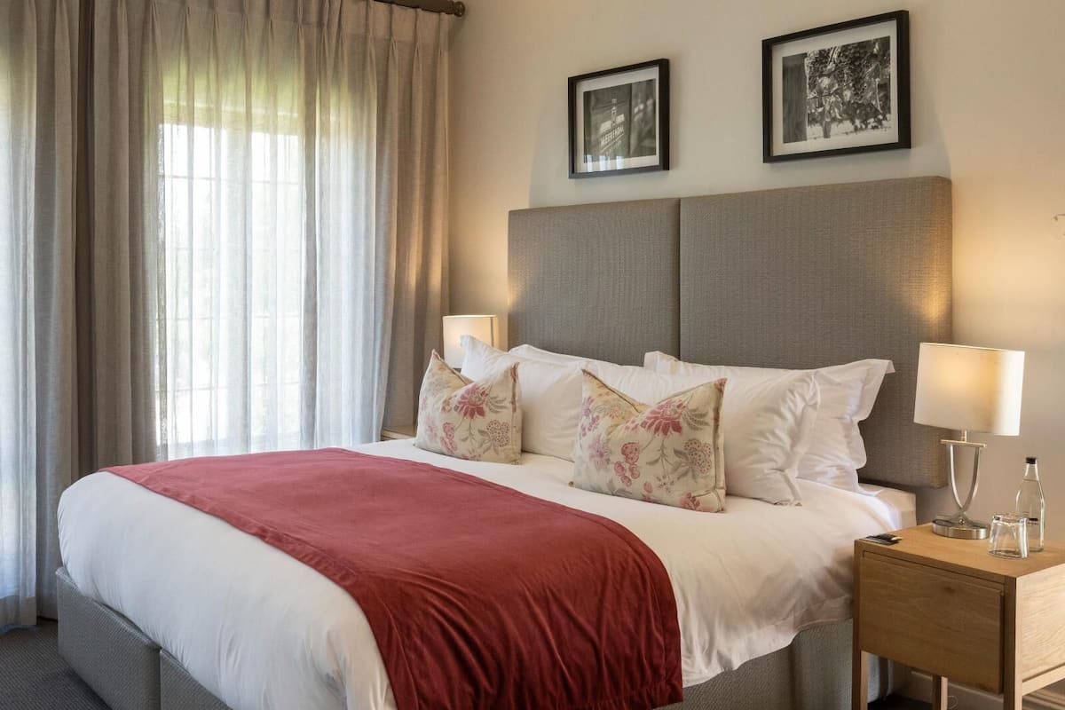 A neatly made bed with a red blanket, two pillows, and a wooden nightstand with a lamp and a bottle.