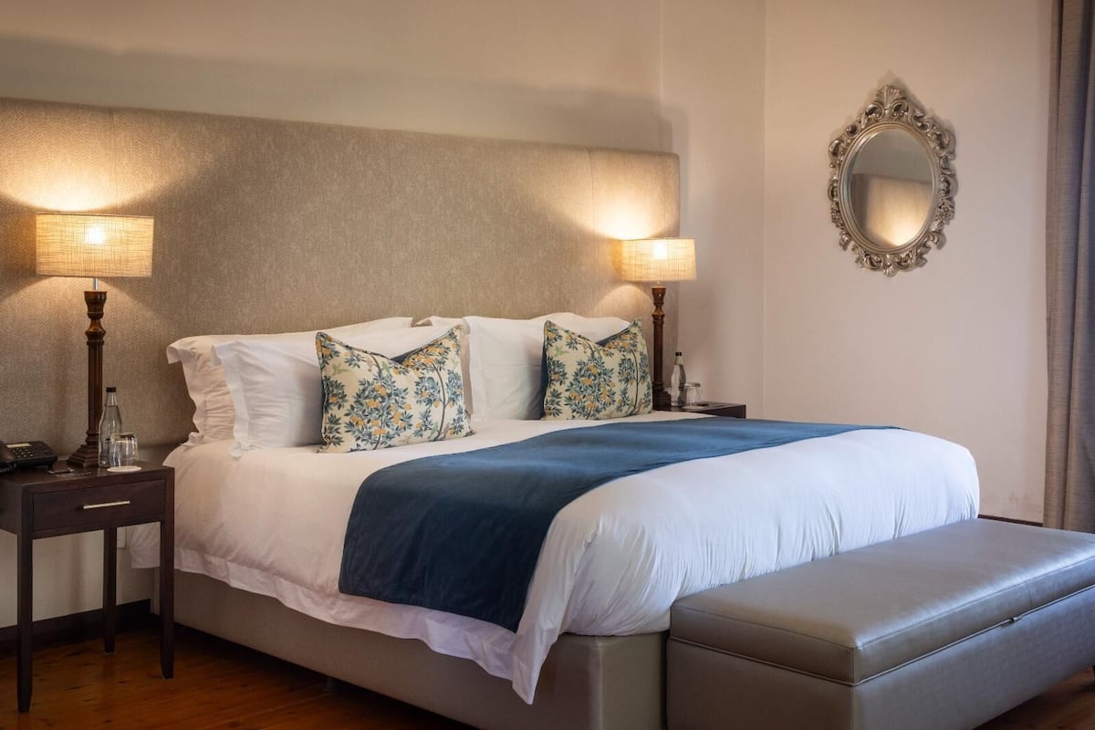 A neatly made bed with a blue blanket, two bedside lamps, a mirror, and a wooden nightstand with a phone and glasses.