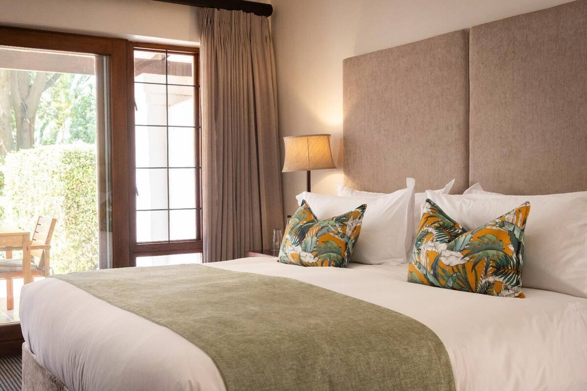 A hotel room with a bed, a window with curtains, a lamp, and a view of greenery outside.