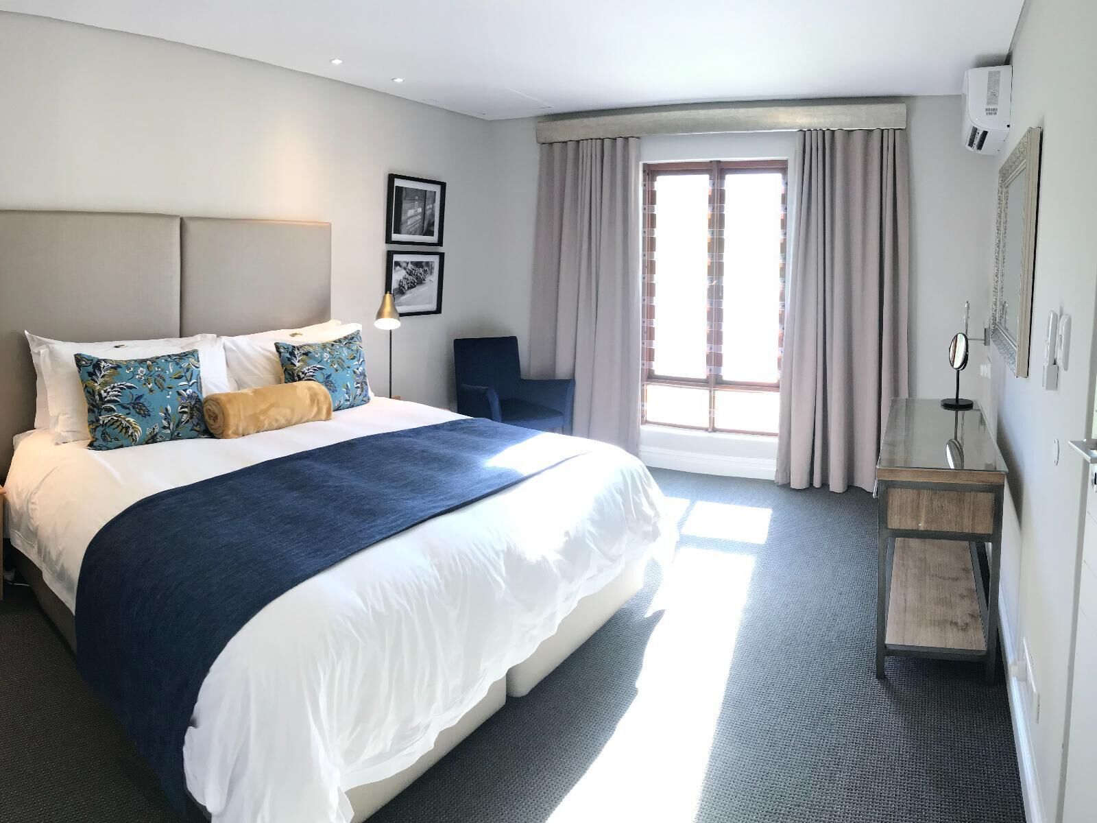 A hotel room with a large bed, a desk, a chair, a window with curtains, and a wall-mounted air conditioner.