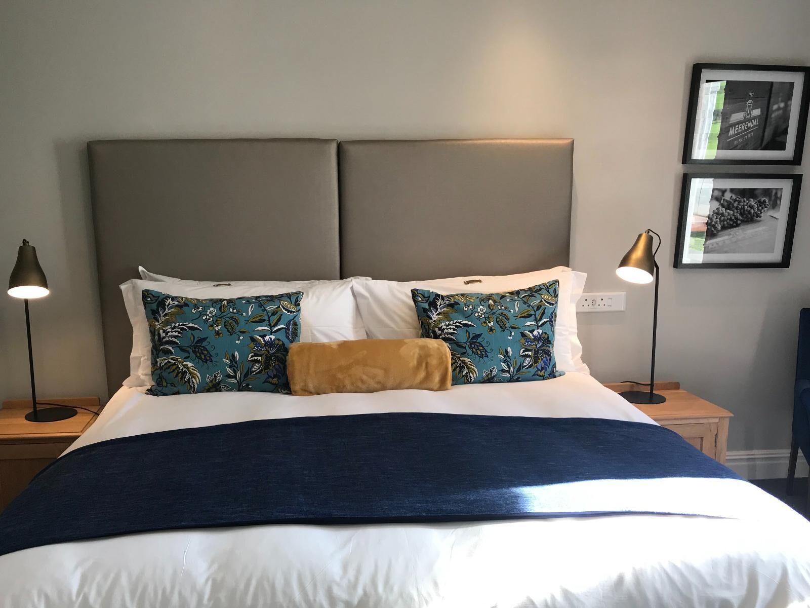 A neatly made bed with a blue and white duvet, two pillows with a floral pattern, and a tan throw. There are two bedside lamps, a framed picture on the wall, and a small wooden nightstand.