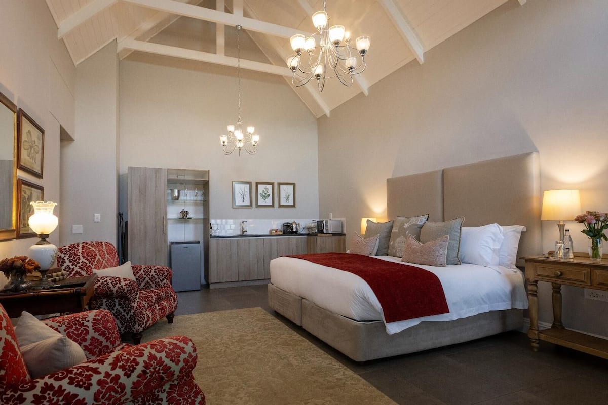 A bedroom with a large bed, a sitting area with patterned chairs, a kitchenette, and a chandelier.
