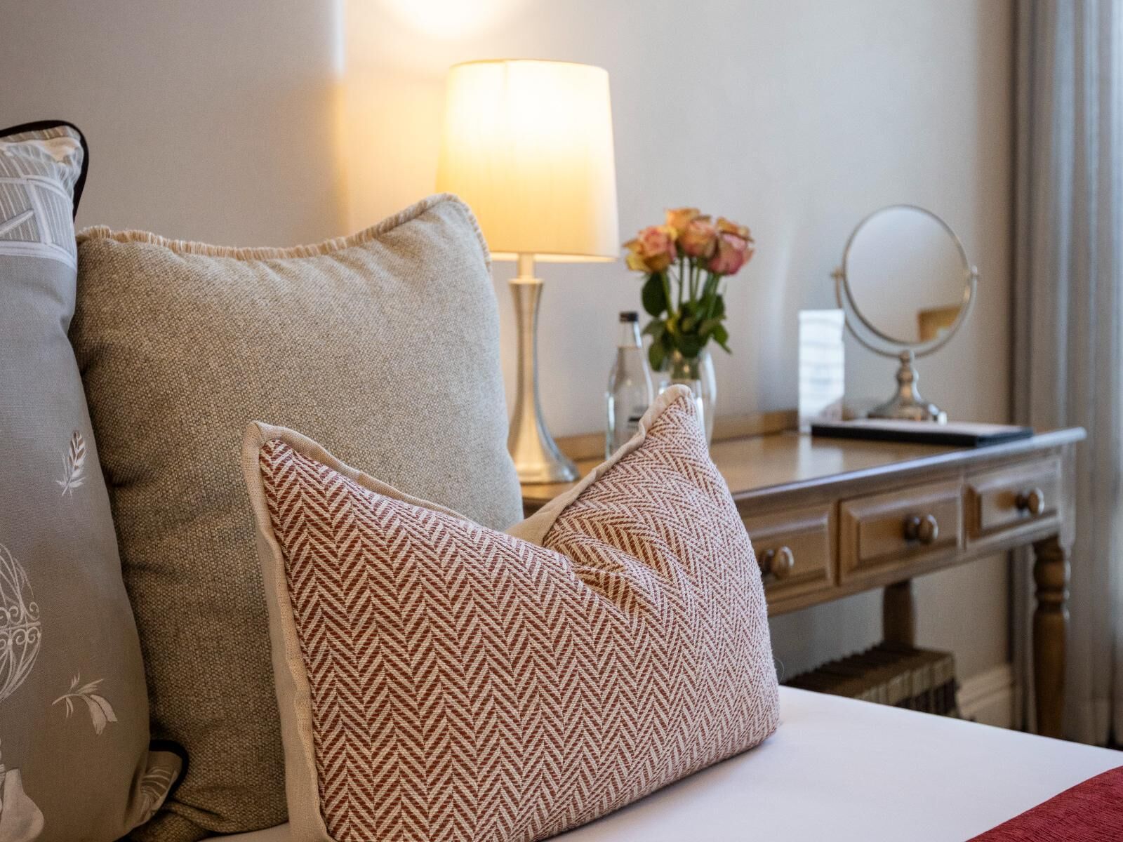 Un lit bien fait, avec deux oreillers, une lampe de chevet, un vase rempli de fleurs et un miroir posé sur une commode.