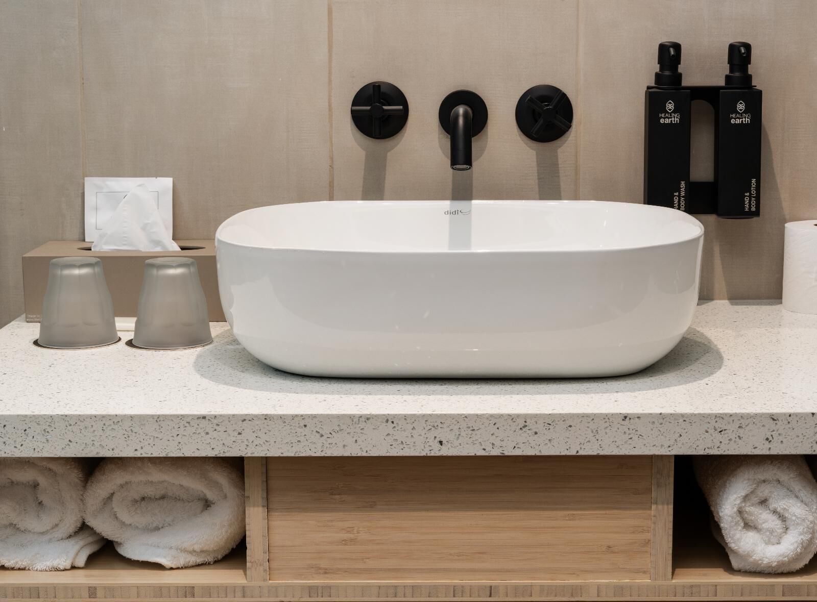 A bathroom with a white vessel sink, two black soap dispensers, a tissue box, and two rolled towels.