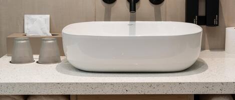 A bathroom with a white vessel sink, two black soap dispensers, a tissue box, and two rolled towels.