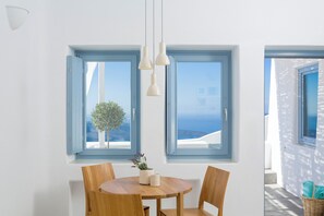 A dining area with a round wooden table and chairs, two windows with blue frames, and hanging pendant lights. - Helestia Pocket Hotel, Caldera Santorini (Santorini)