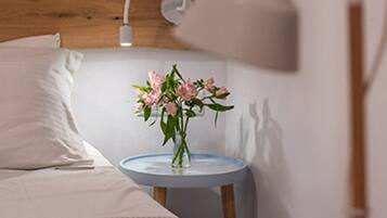 Una cama bien hecha con sábanas blancas, una mesita de noche con un jarrón de flores rosadas, una lámpara blanca y un cabecero de madera.