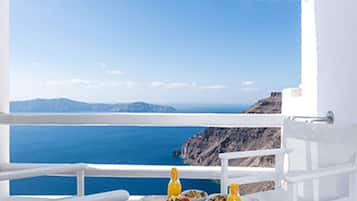 Varanda branca com mesa e cadeiras, com vista para um mar azul límpido e montanhas ao longe.