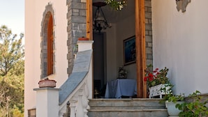 A stone staircase leading to a wooden door with a hanging wreath, a blue mat with "PANTHEON" on it, and potted plants on the steps. - Pantheon Studios Apartments (Thasos)