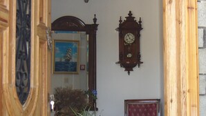 A room with a wooden door, a blue ceramic pot with plants, a red upholstered chair, a wall-mounted clock, and a chandelier. - Pantheon Studios Apartments (Thasos)