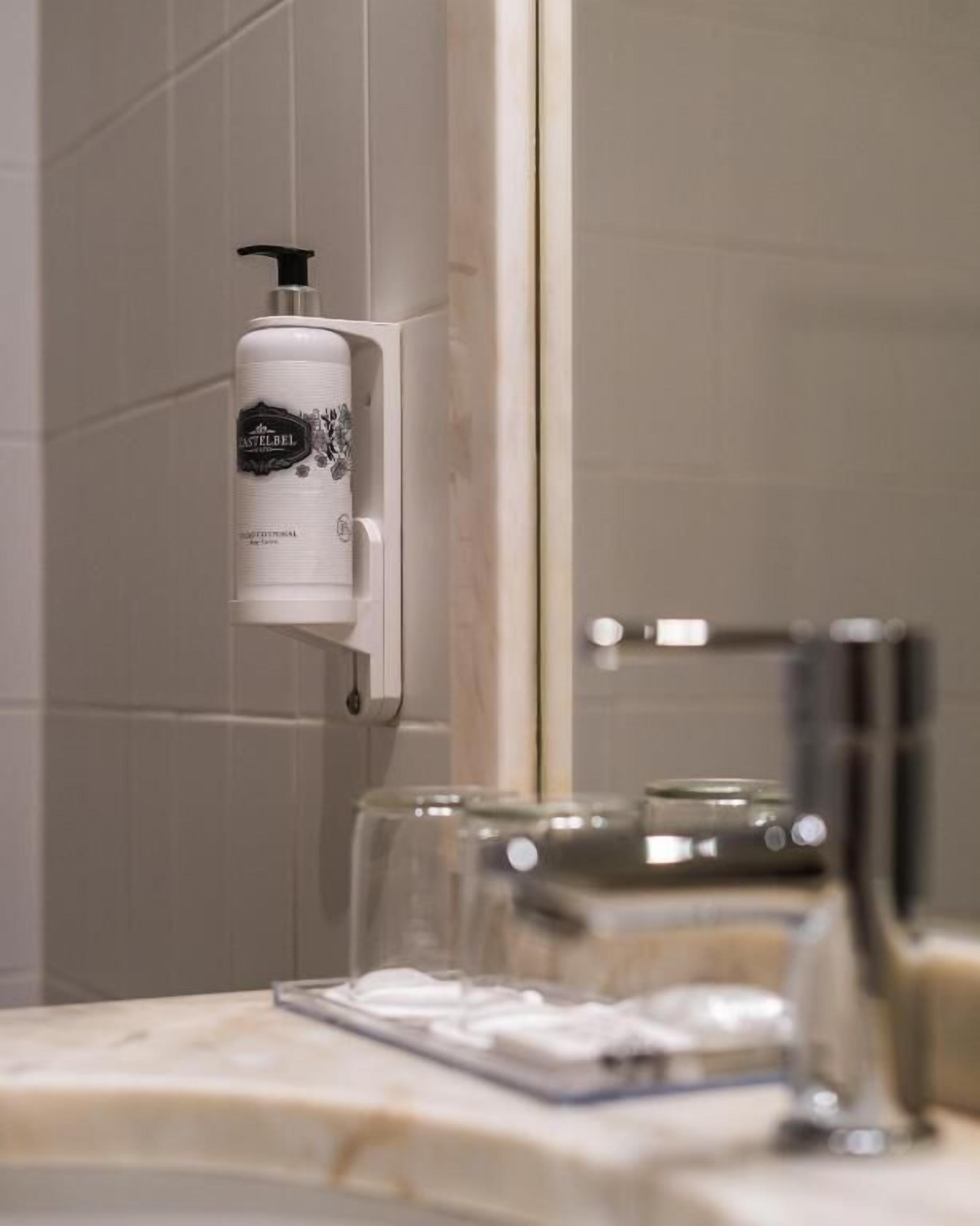 Un baño con dispensador de jabón en la pared, un espejo y un lavamanos con grifo.