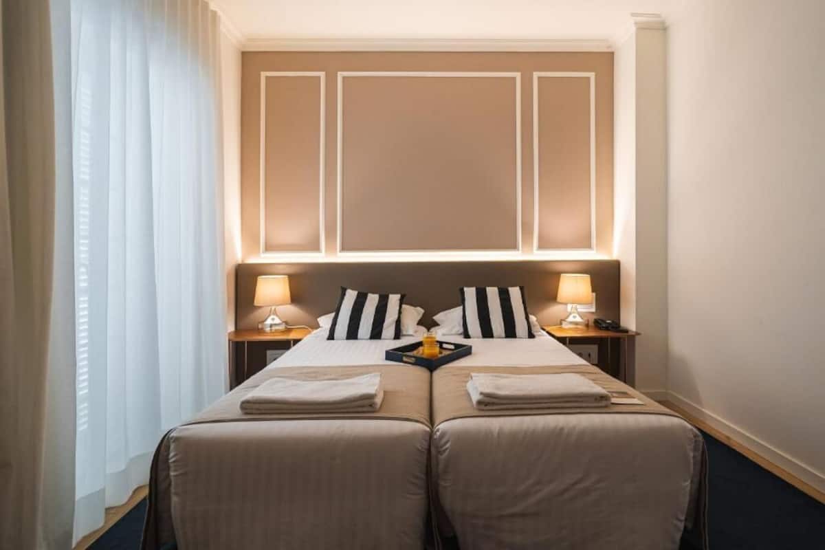 Une chambre d’hôtel avec deux lits, une tête de lit dotée d’un éclairage intégré, des tables de chevet avec des lampes et une fenêtre avec des rideaux.