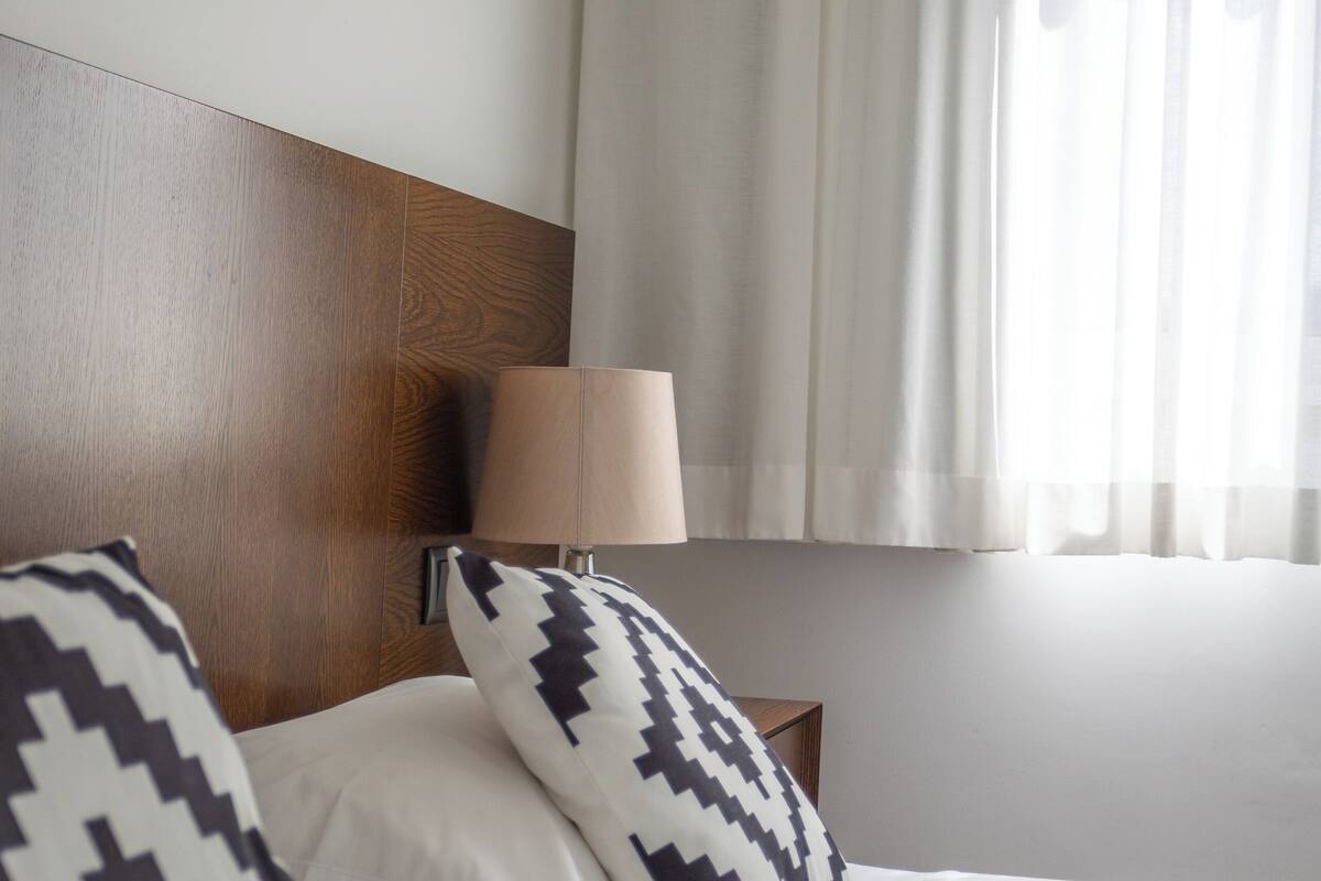 Une chambre d’hôtel avec deux lits, une tête de lit en bois, une table de chevet avec une lampe et des rideaux légers.
