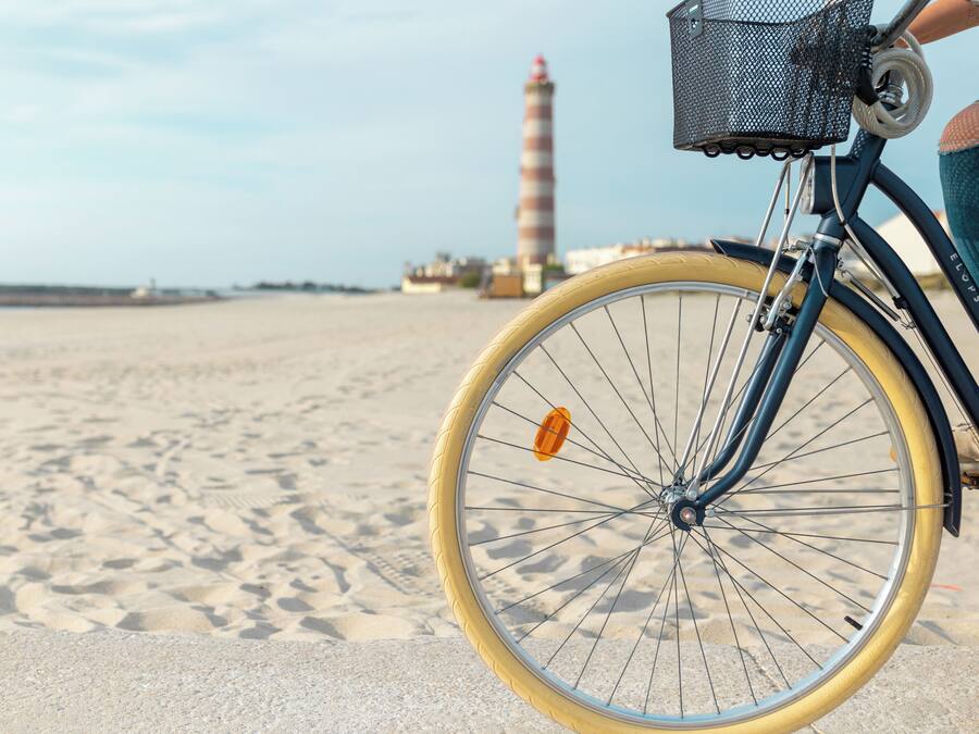Een fiets met een mandje op een zanderig strand, met een vuurtoren op de achtergrond.