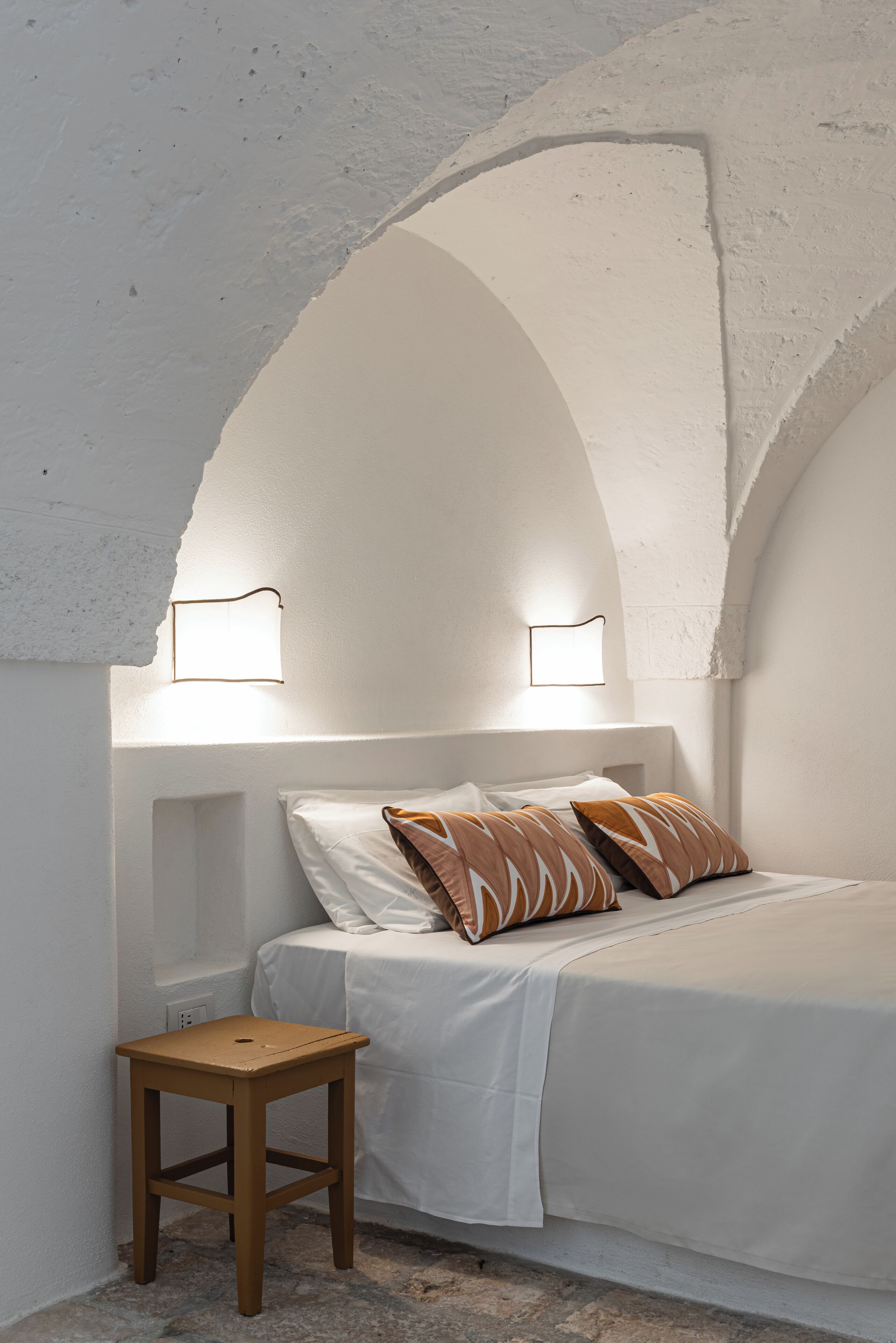 Un dormitorio con una cama blanca, dos almohadas estampadas, un taburete de madera y dos luces fijadas a la pared.