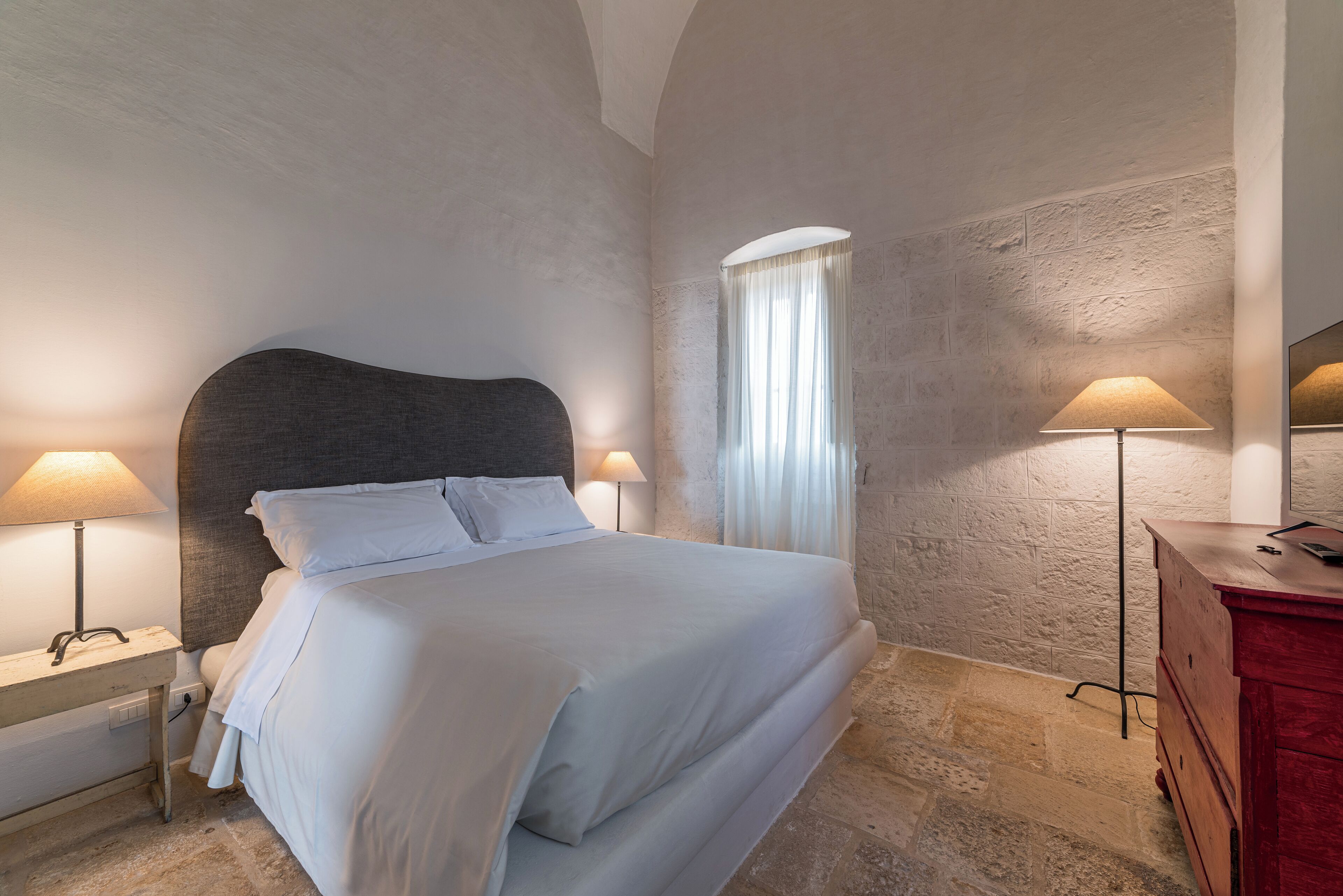 Ein Schlafzimmer mit einem großen Bett, zwei Nachttischlampen, einem Nachttisch, einer Kommode und einem Fenster mit Vorhängen.