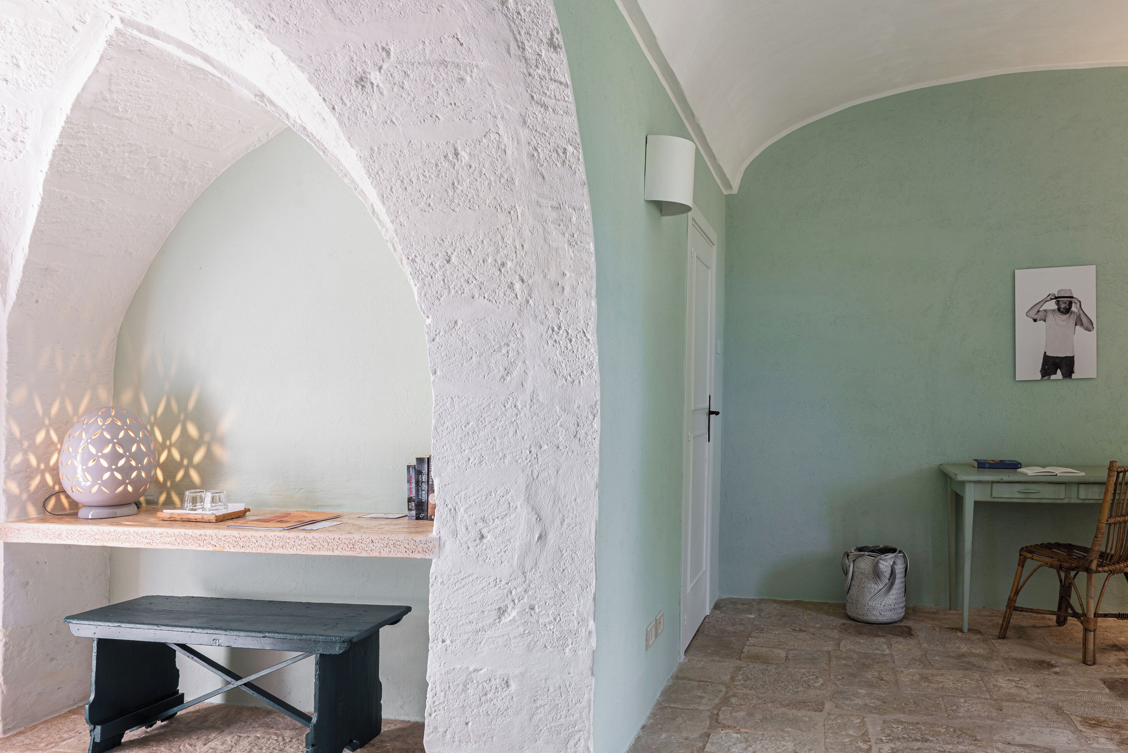 A room with a stone floor, a desk with a lamp, a chair, and a wall with a framed picture.