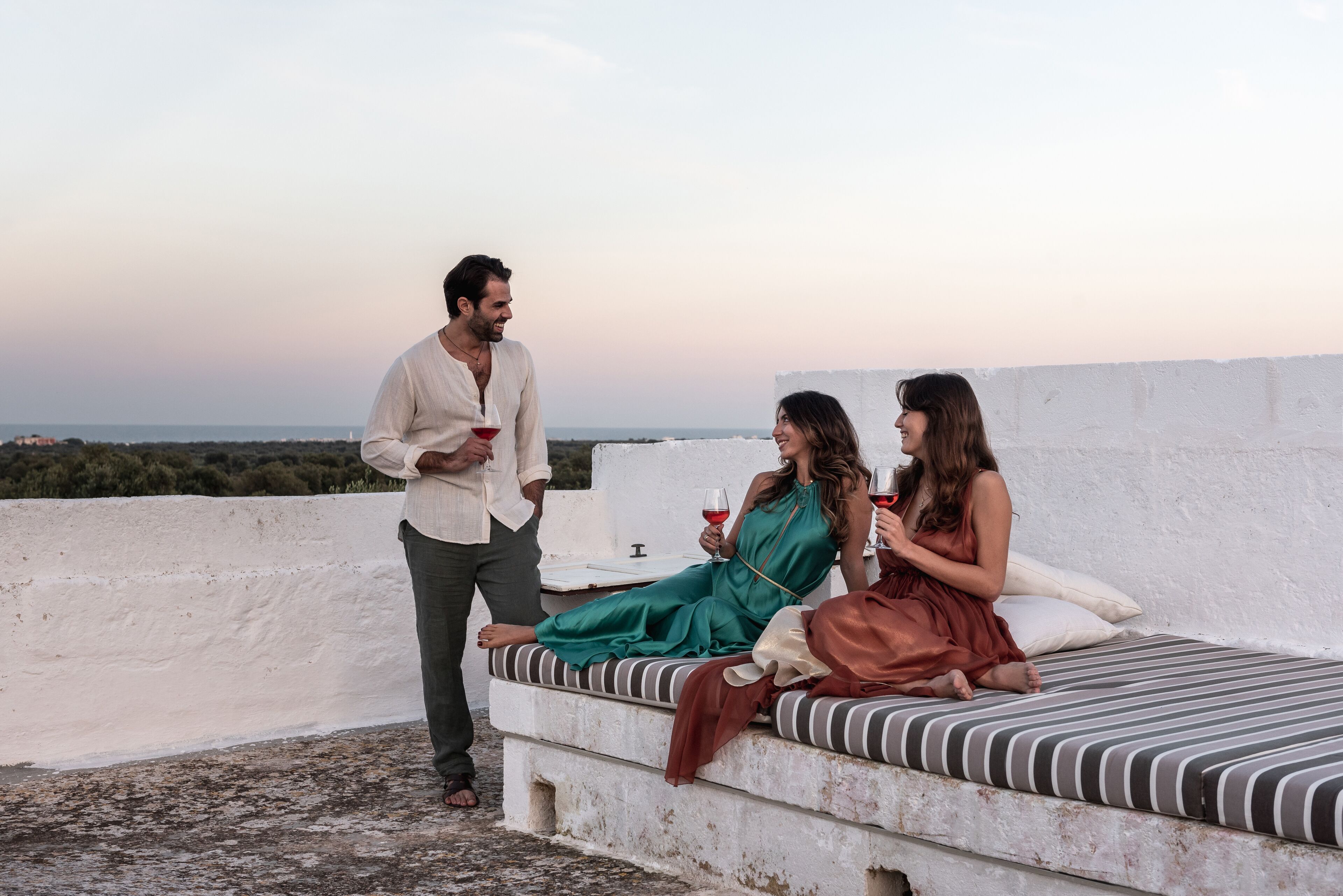 A rooftop terrace with a bed, a man holding a drink, and two women sitting.