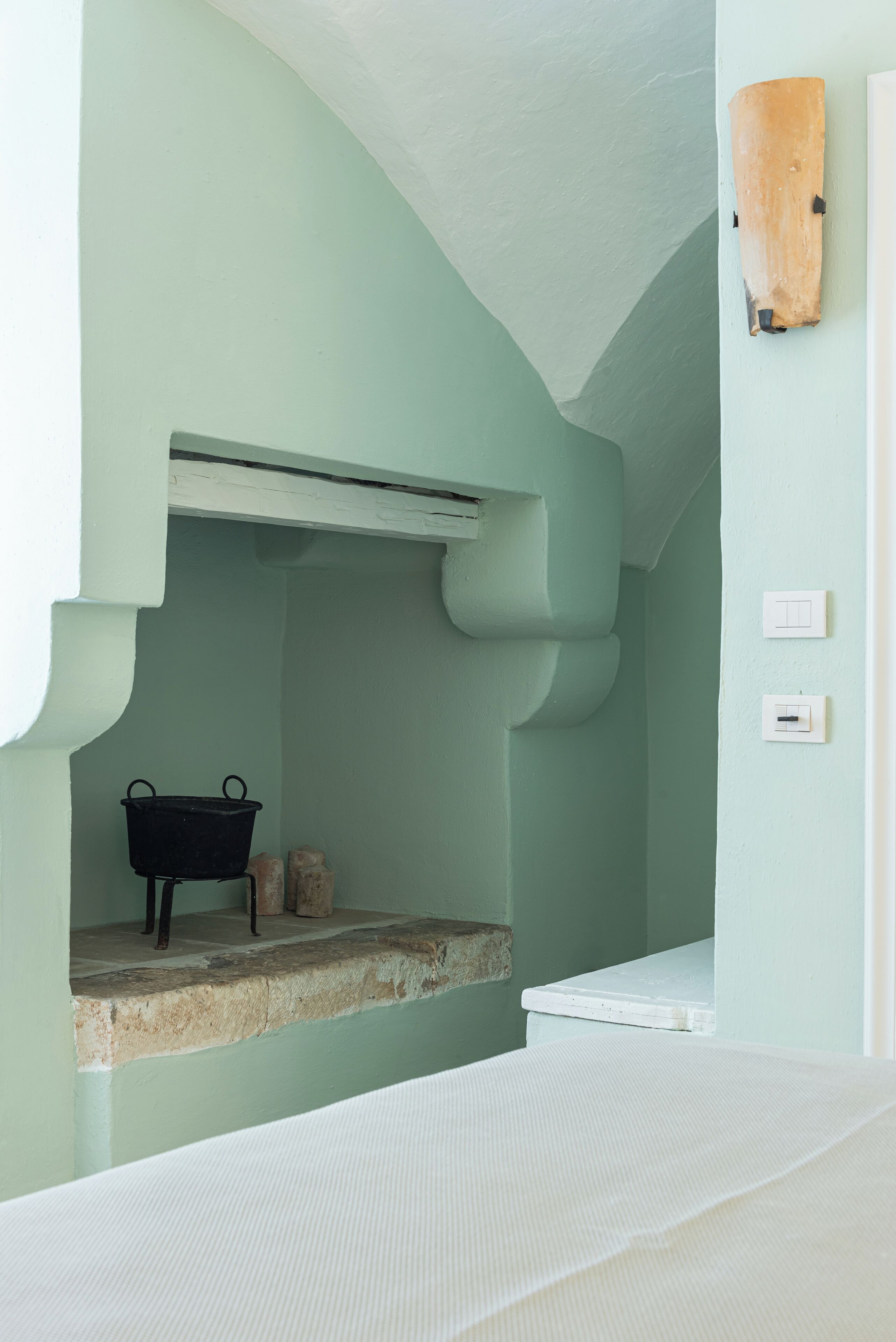 Una habitación con techo bajo, una cama blanca, un estante pequeño con una olla negra y un interruptor de luz en la pared.