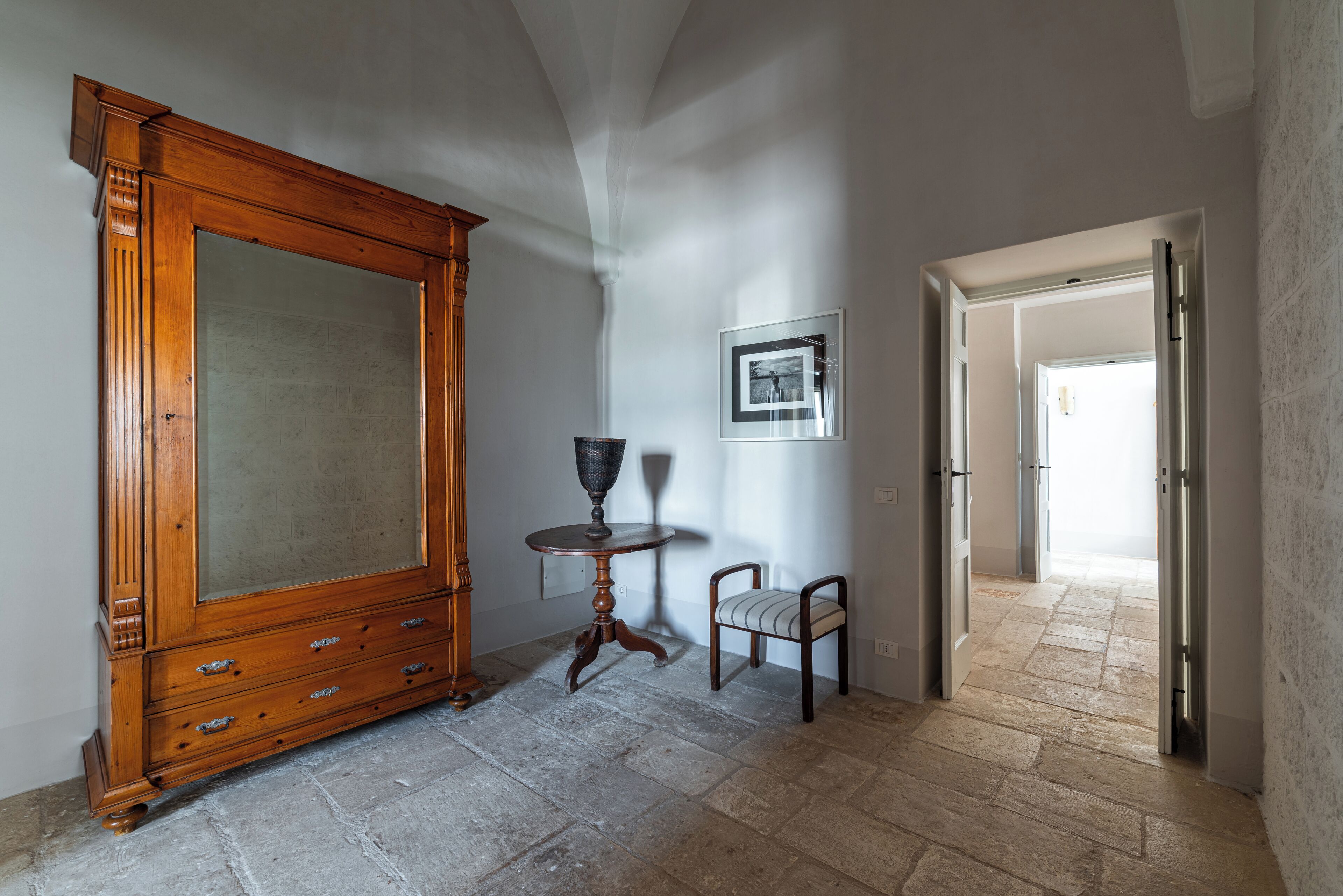 A room with a large wooden cabinet, a small round table, a chair, and a framed picture on the wall.