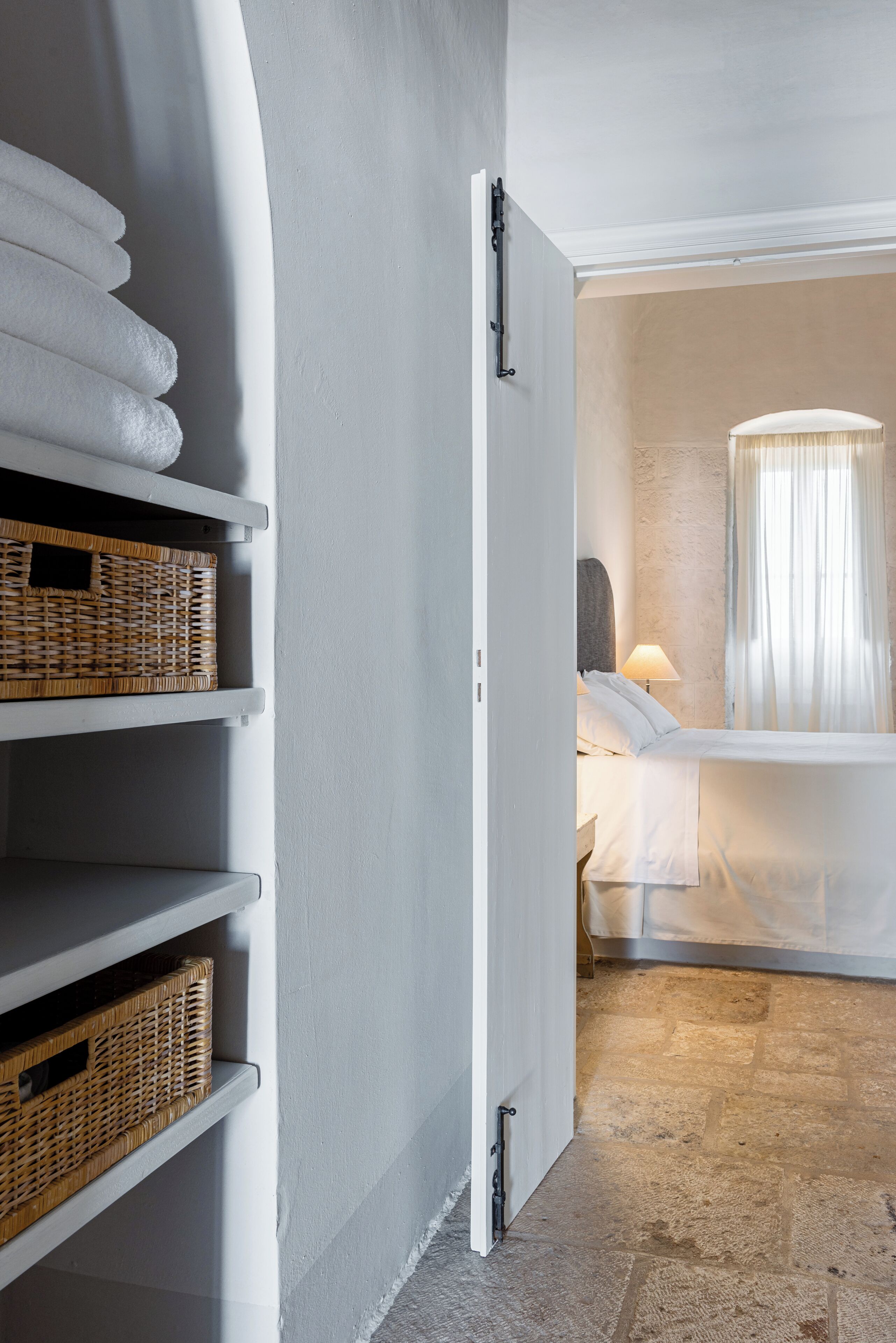 A hotel room with a bed, a nightstand, a lamp, and wicker baskets on shelves.