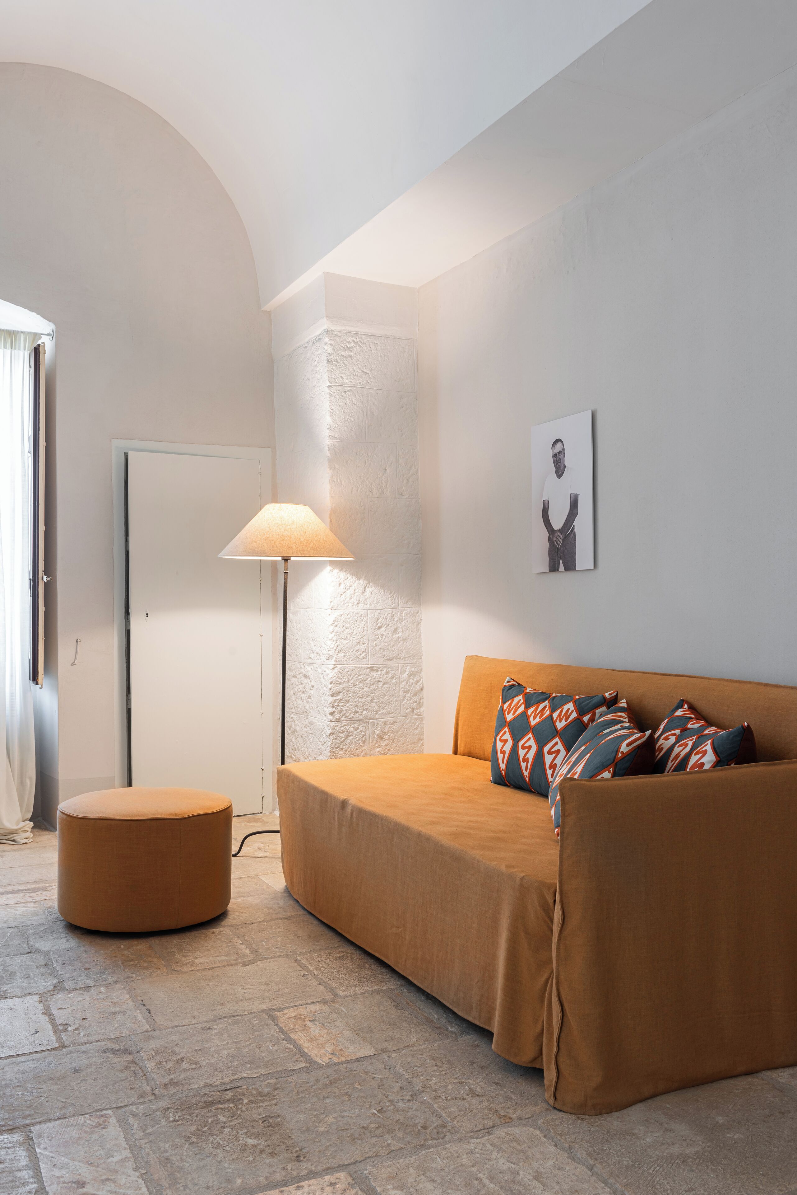 A minimalist living room with a brown sofa, patterned cushions, a round ottoman, a floor lamp, and a framed picture on the wall.