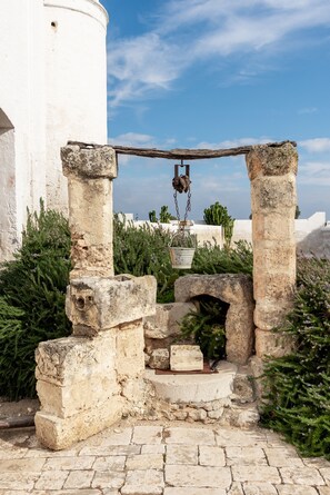 Una antigua estructura de piedra con una olla y cadena colgantes, rodeada de follaje y un edificio blanco al fondo.
