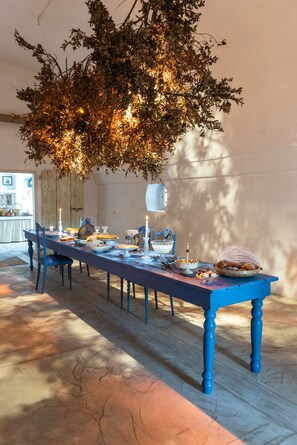 Una larga mesa de comedor con sillas azules, adornada con velas y platos, bajo un árbol grande.