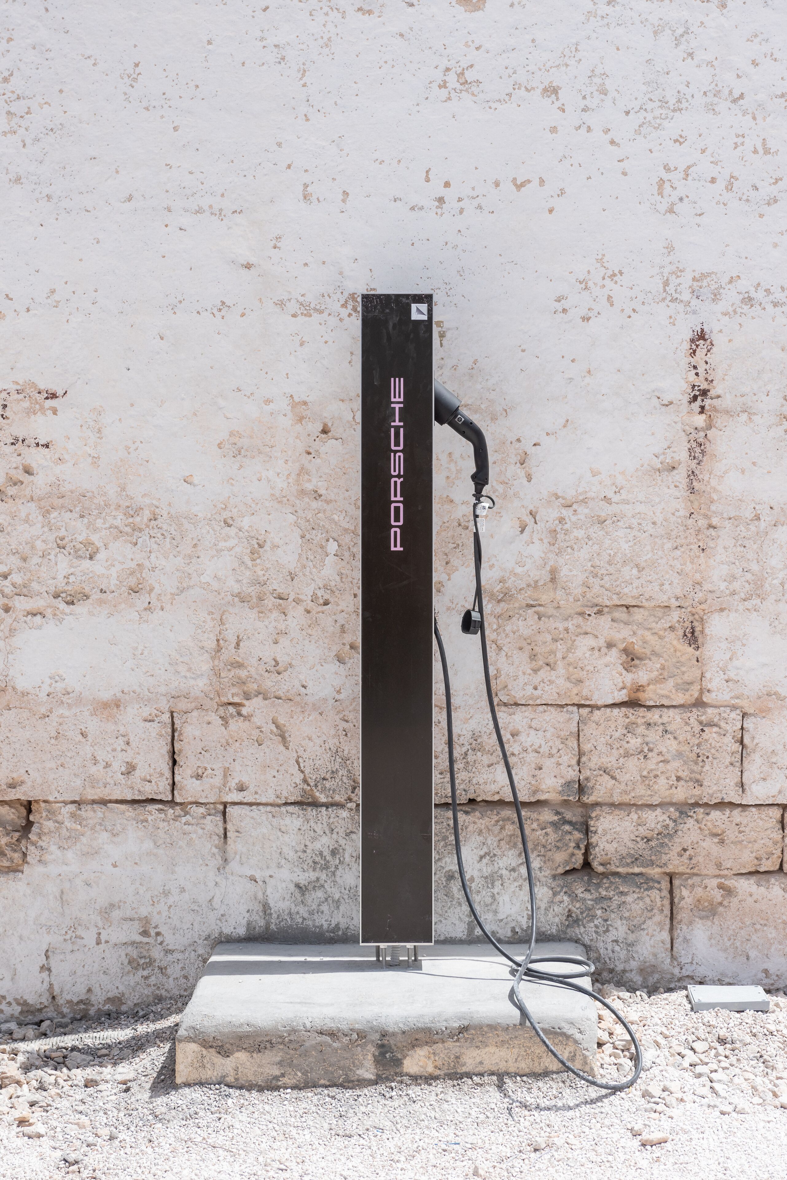 A black electric vehicle charging station with the Porsche logo, mounted on a concrete base against a textured wall.