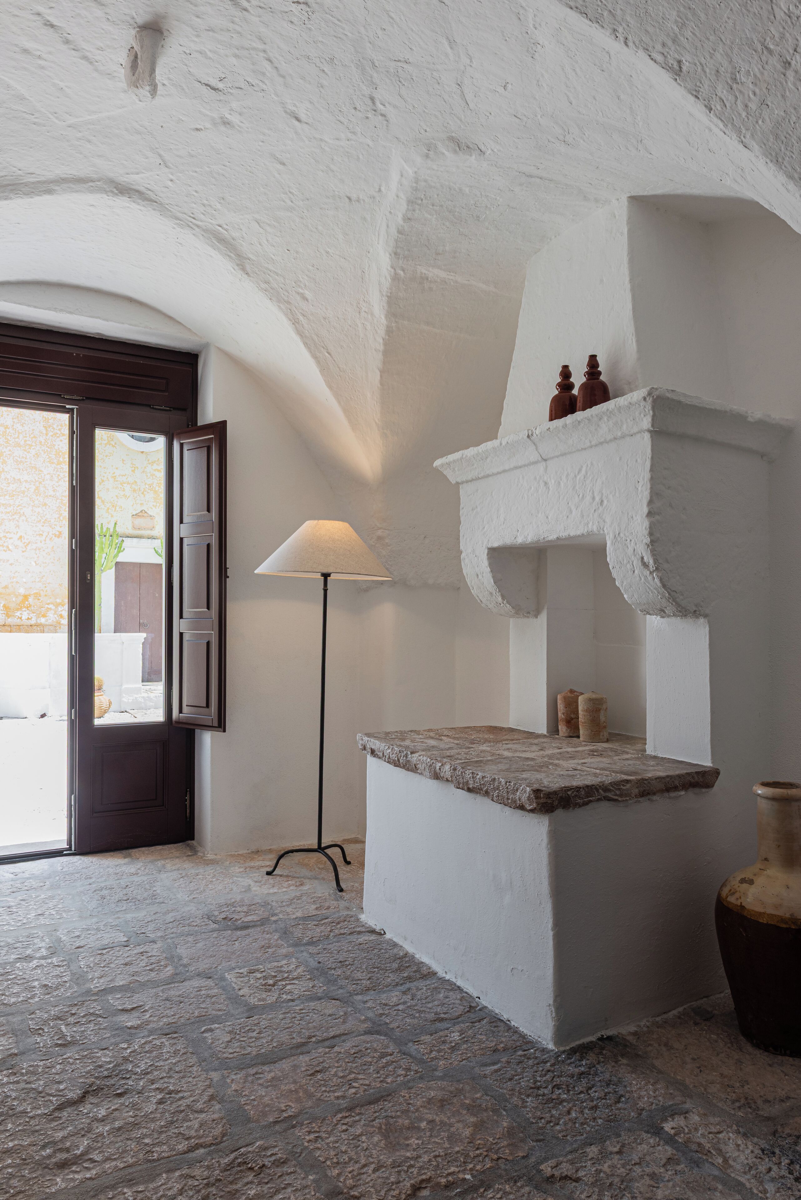 A rustic interior with a stone floor, a white wall with a shelf, a lamp, and a door leading outside.