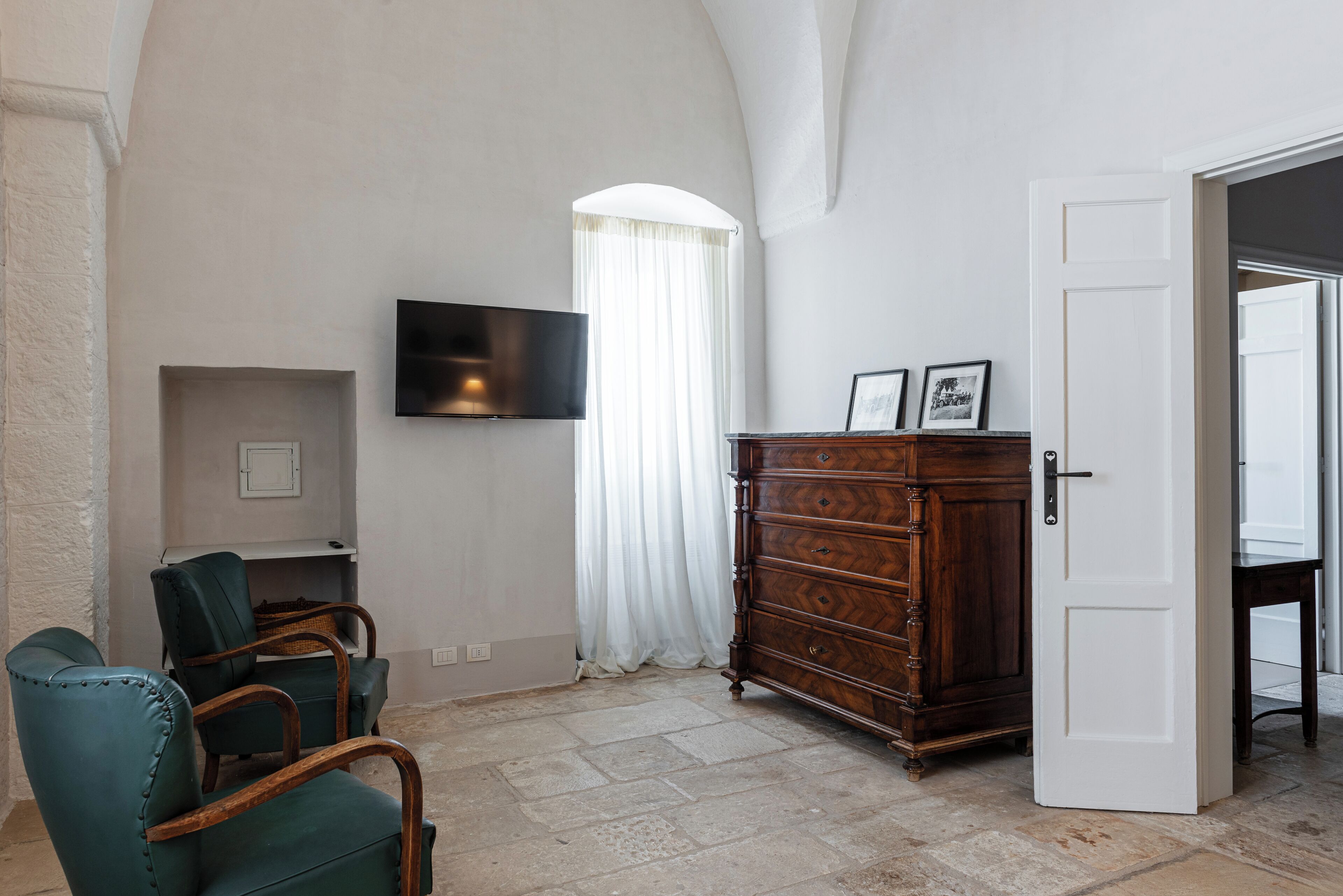 Une pièce dotée d’un téléviseur à écran plat fixé au mur, d’une commode en bois avec des tiroirs, d’un fauteuil vert recouvert de tissu et d’une porte blanche donnant sur une autre pièce.