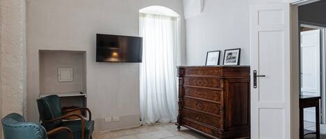 Une pièce dotée d’un téléviseur à écran plat fixé au mur, d’une commode en bois avec des tiroirs, d’un fauteuil vert recouvert de tissu et d’une porte blanche donnant sur une autre pièce.