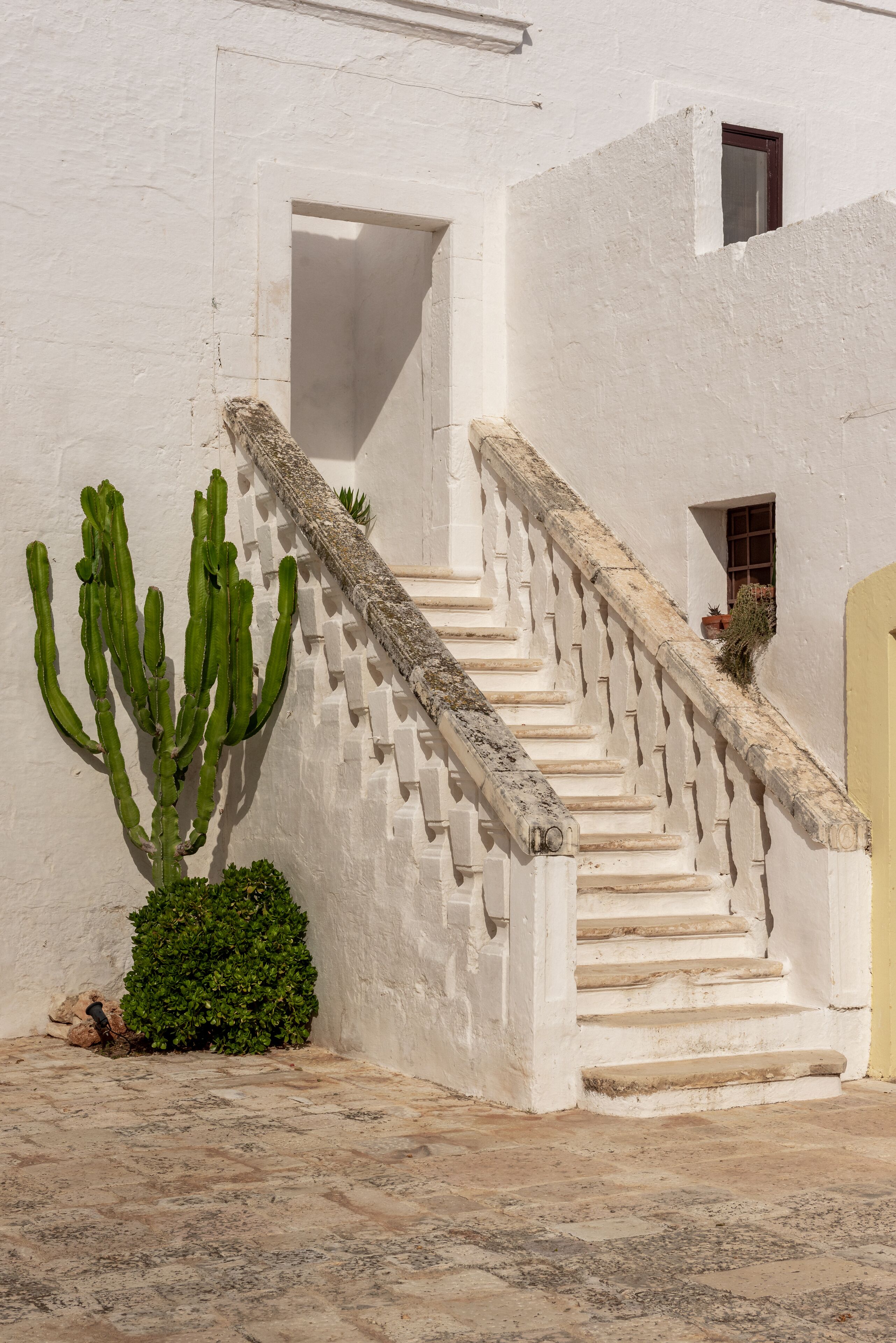 A white building with a staircase leading to a door, a cactus, and a small window.