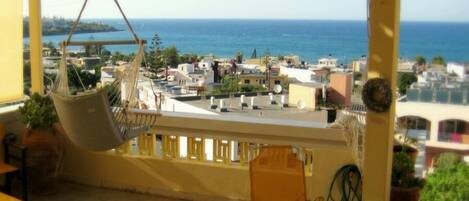 A balcony with a hanging chair, potted plants, and a view of the sea and buildings.