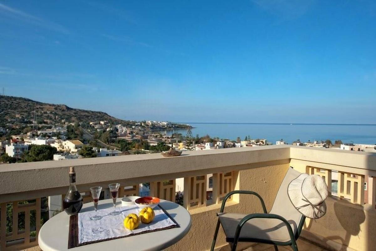 A balcony with a table set for two, overlooking a coastal town and the sea.