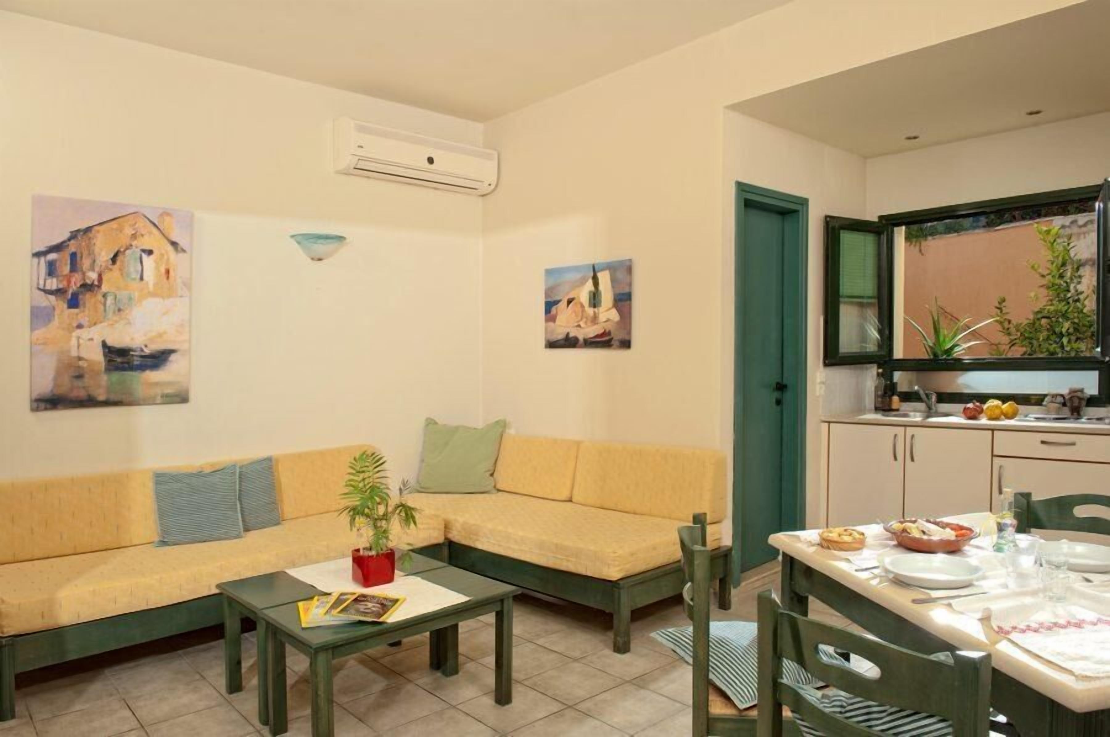 A living room with a sofa, a coffee table, and a dining area with a table set for two. There are paintings on the walls and a kitchen area with cabinets and a sink.