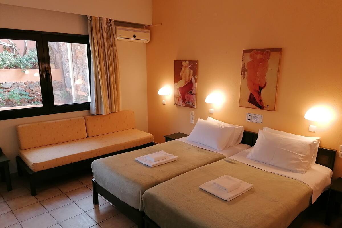 A hotel room with two beds, a sofa, a window with curtains, and wall-mounted lights.