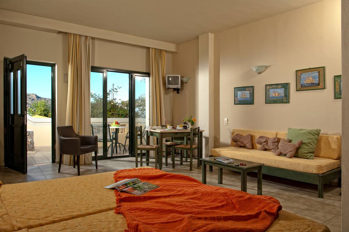 A hotel room with a bed, a sofa, a dining table with chairs, a TV, and a balcony with curtains.