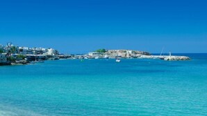 A coastal town with buildings along the shore, a rocky island with a lighthouse, and boats in the water.