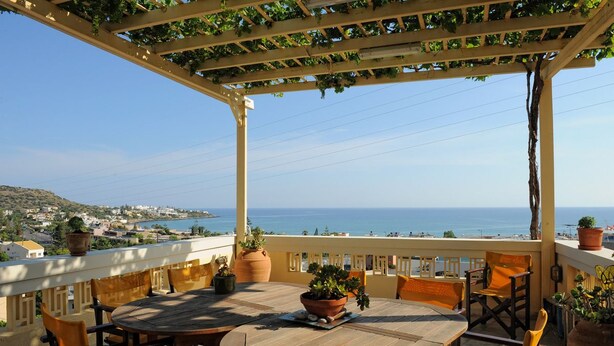A terrace with a wooden table and chairs, potted plants, and a view of the sea.