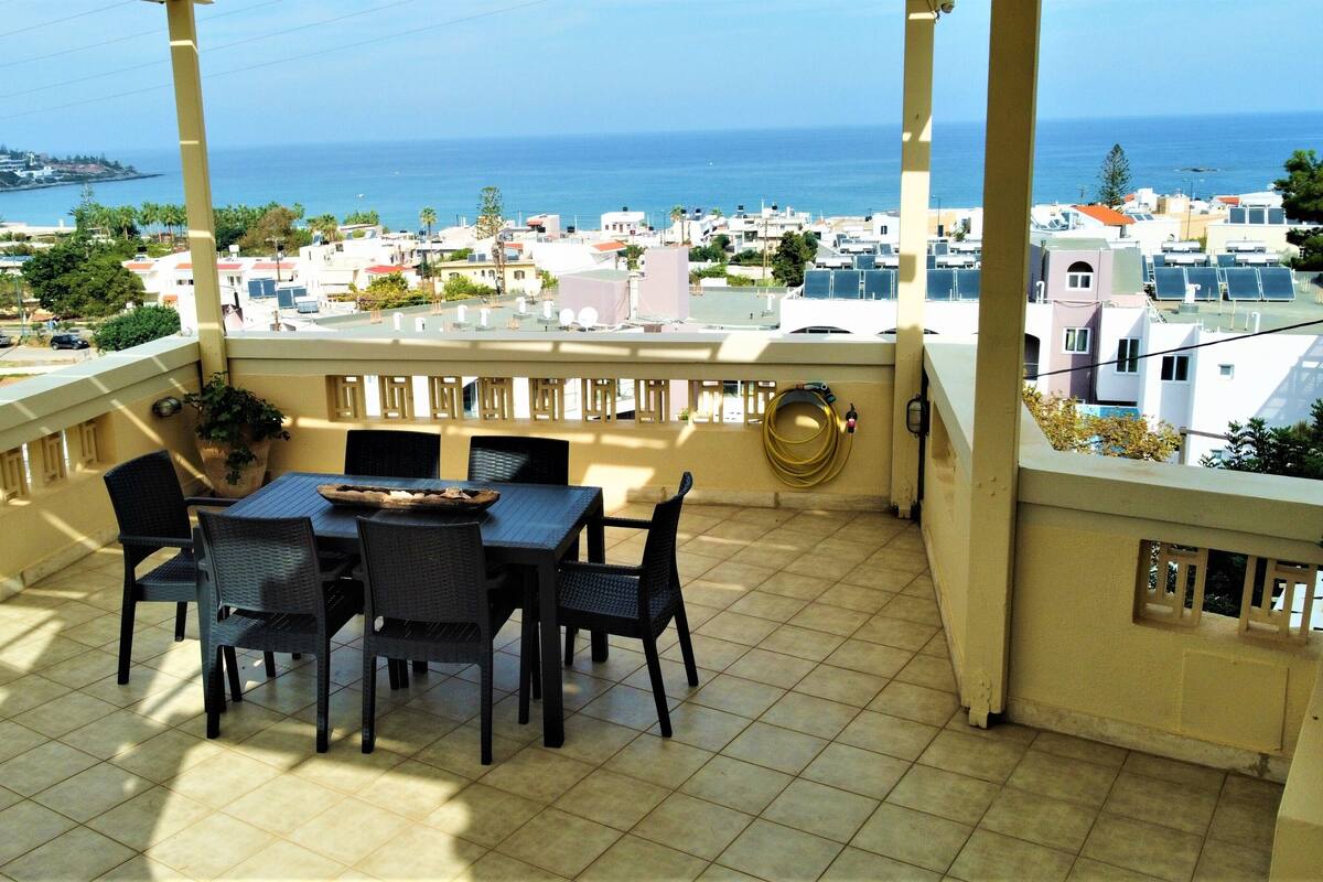 A balcony with a table and chairs, overlooking a coastal town with buildings and the sea.