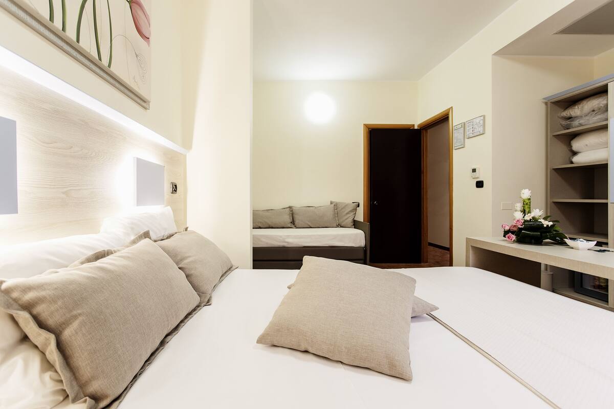 Une chambre d’hôtel avec un grand lit, un lit plus petit, un bureau agrémenté d’un vase de fleurs et une armoire.