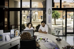 Una oficina con escritorio, computadora y varios documentos. Hay ventanas con vista a la calle y edificios.