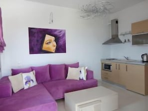 Ein modernes Wohnzimmer mit einem violetten Sofa, einem weißen Couchtisch und einer Küchenzeile mit Schränken und einer Mikrowelle.