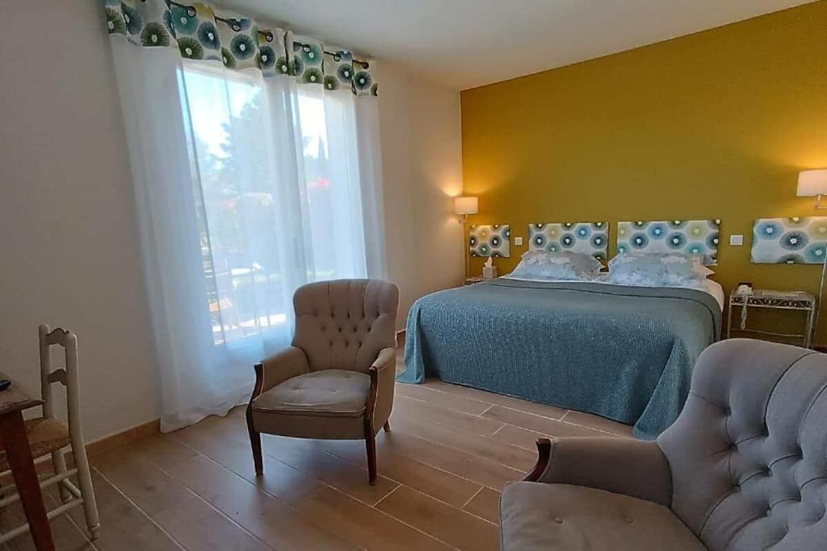 A bedroom with a bed, two armchairs, a wooden chair, a nightstand, and a window with curtains.