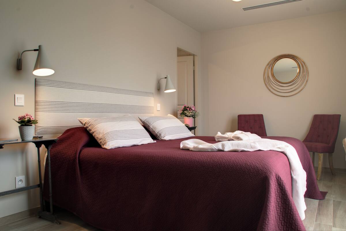 A neatly made bed with a striped headboard, a round mirror on the wall, and a red chair.