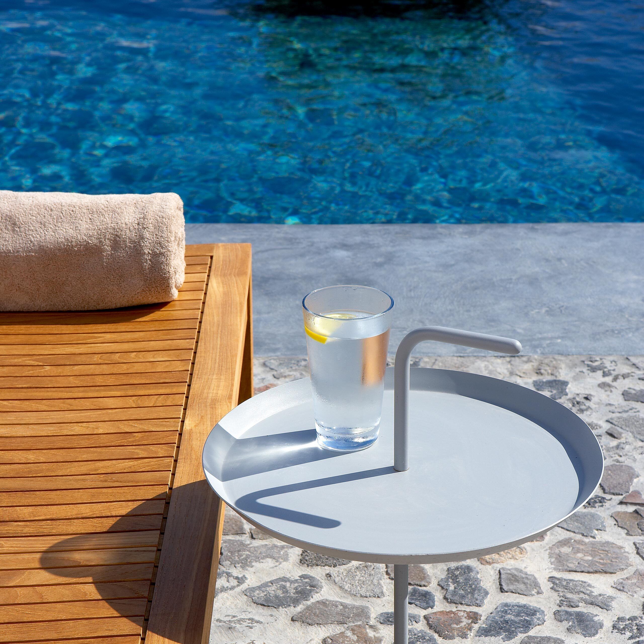 Ein Poolbereich mit einer Holzliege, einem Glas Wasser mit Zitronenscheibe und einem Metalltisch mit weißem Teller.
