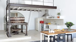 A modern kitchen with a rustic wood oven, a dining area with a wooden table, and a loft space above. - Villa Fabrica Santorini (Santorini)